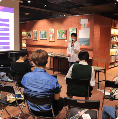 교육 사업부 강의 현장 이미지, 한 남성이 마이크를 들고 강의 중이며 세 명의 사람이 의자에 앉아 강의를 듣고 있다